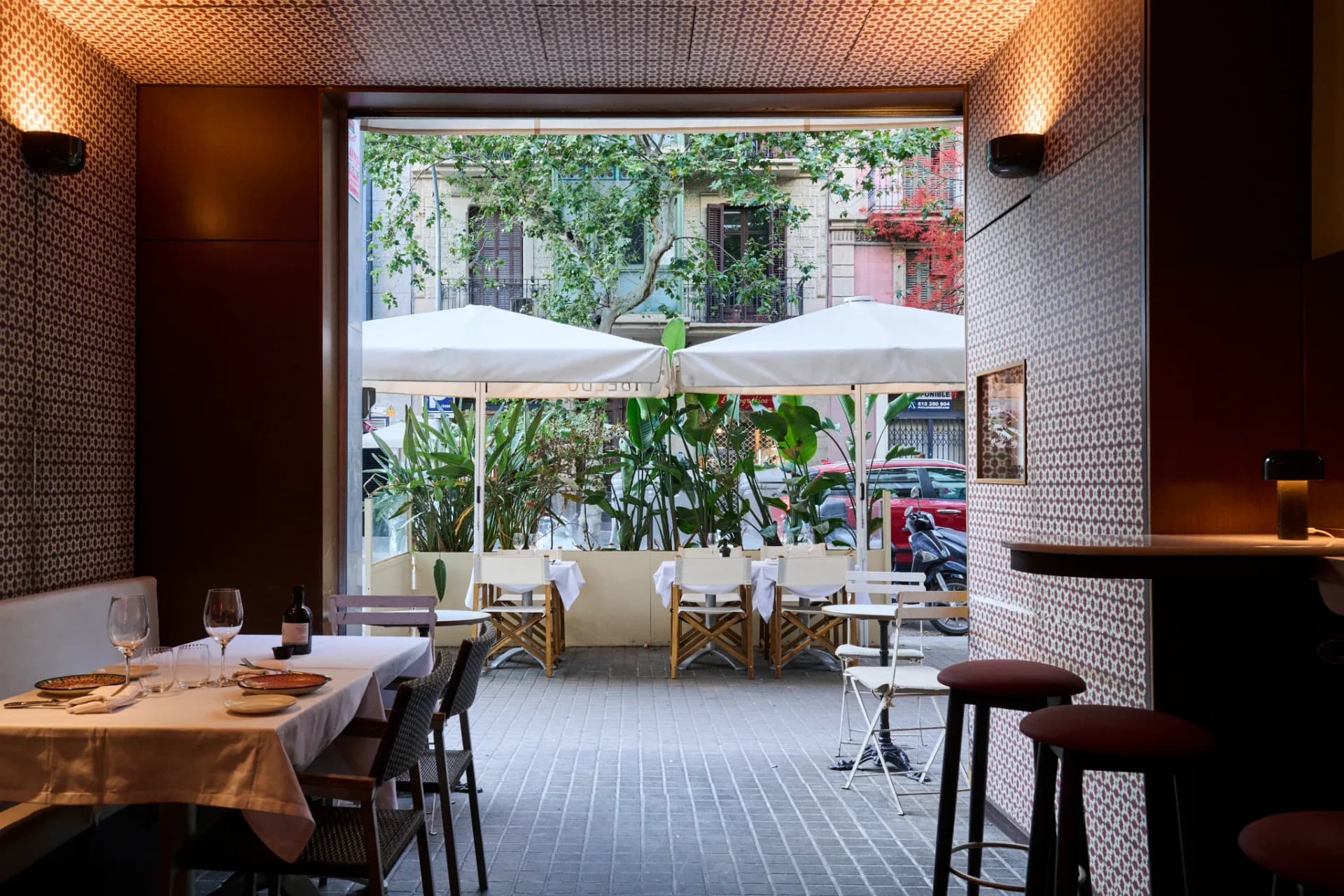 Vista desde el interior del Restaurante Igueldo en Barcelona hacia la terraza con mesas, sillas de madera y plantas junto a la calle